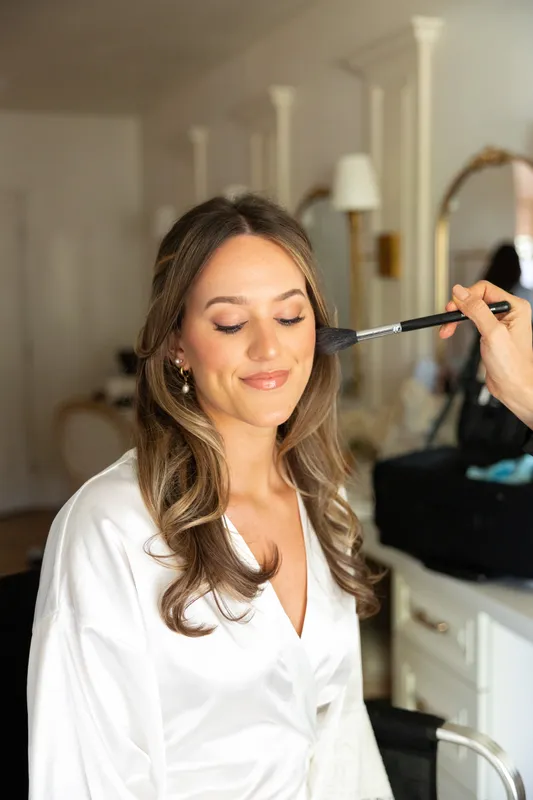 Makeup artist using a brush to apply blush on a bride with long wavy hair wearing a white robe.