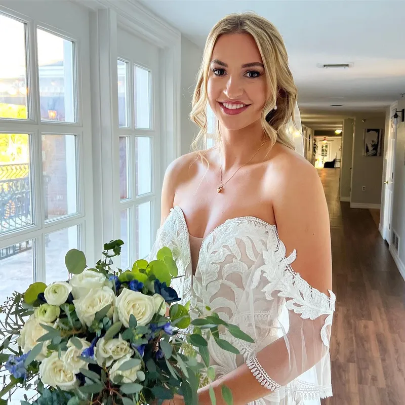 A beautiful bride with elegant professional makeup holding a white floral wedding bouquet by a window.