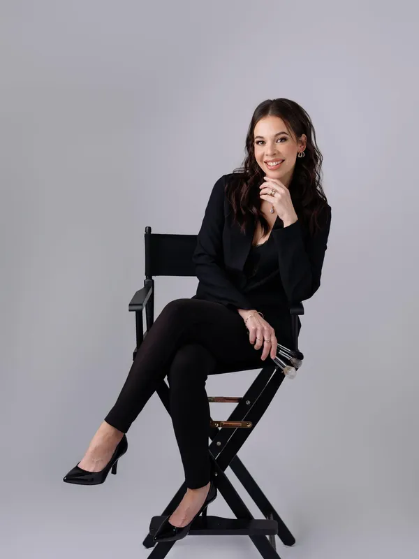 A professional makeup artist sitting in a director chair holding makeup brushes against a grey background.