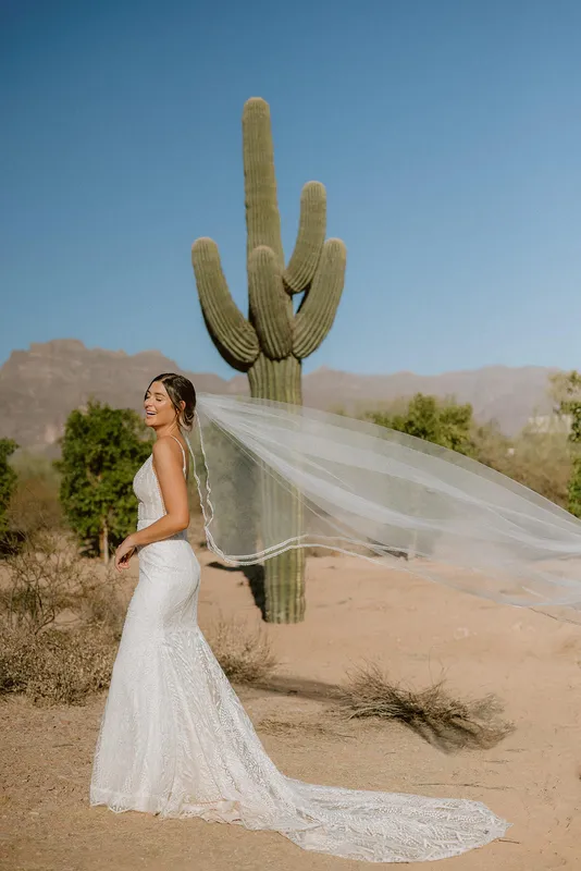 Bride with elegant wedding makeup and a flowing veil posing in the Arizona desert near a Saguaro cactus.