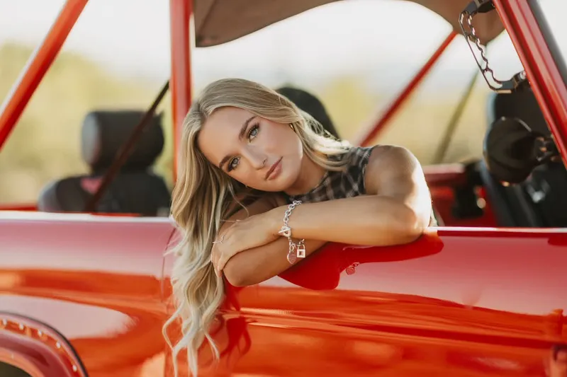 Young woman with professional senior portrait makeup leaning on a bright red vintage car during a photoshoot.