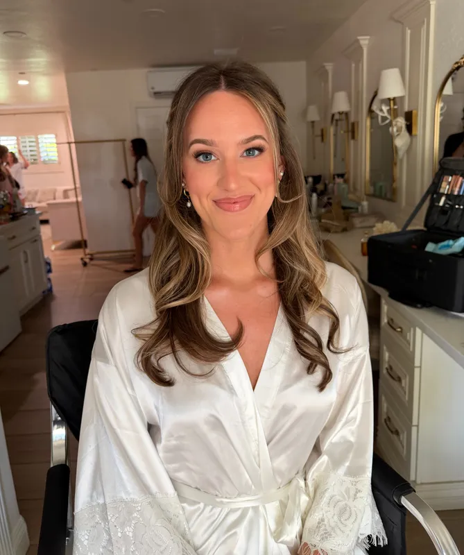 A beautiful bride with soft glam makeup and a white silk robe smiling at the camera.